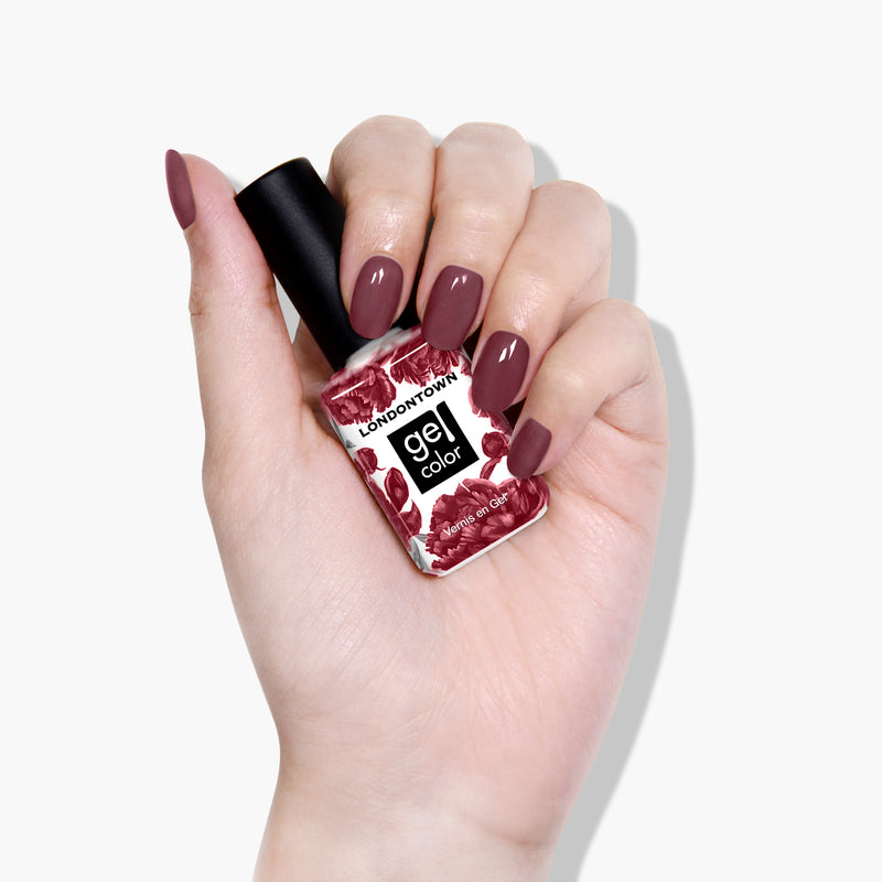A hand with painted fingernails in a rich and ripened mauve-tinged purple holds a bottle of LONDONTOWN gel nail polish in the Fig Jam shade.