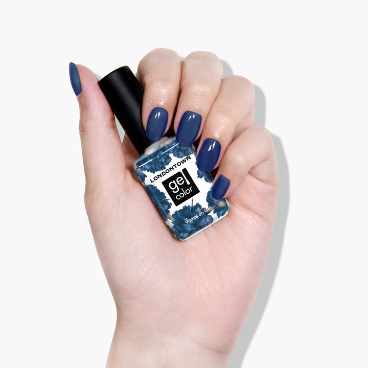 A hand with manicured nails painted in Under The Stars, a vintage denim blue, holds a bottle of LONDONTOWN gel color nail polish.
