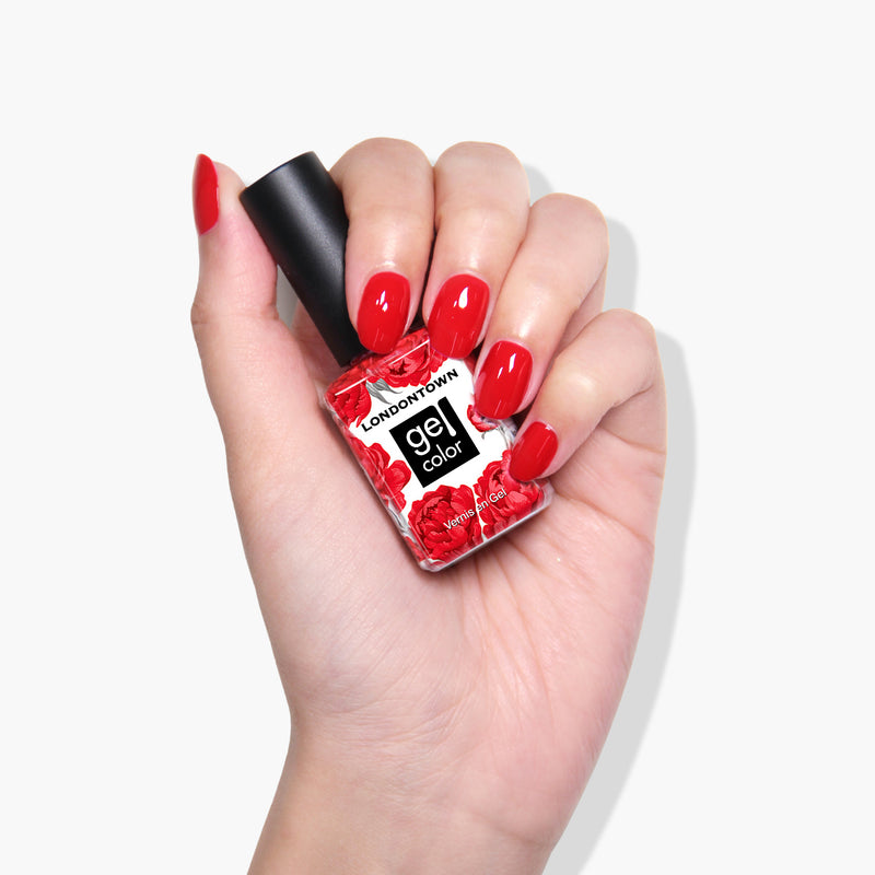 A hand holds a bottle of LONDONTOWN's "Changing of the Guards" gel nail color in deep red crème with red floral designs against a plain white background. The nails, reminiscent of Buckingham Palace elegance, are painted in a matching glossy red shade.