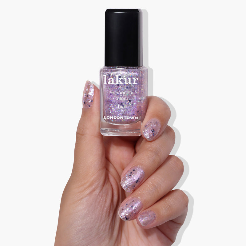 A hand holds a bottle of LONDONTOWN's Grape Fizz Lakur nail polish in a glittery pink shade with hints of holographic purple glitter, while the nails are painted with the same polish.