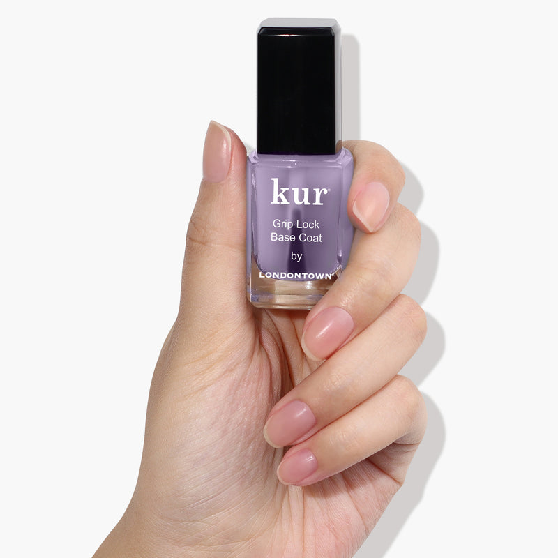 A hand with manicured nails holds a bottle of LONDONTOWN Grip Lock Base Coat nail polish, the superior formula adding extra shine to its light purple color.