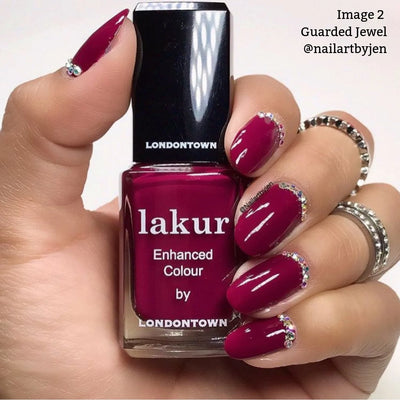 A hand showcases manicured nails featuring a deep maroon crème polish with jeweled accents, clutching a bottle of LONDONTOWN's Guarded Jewel (Professional) enhanced color. Rings embellish the fingers. Text in the corner reads "Image 2 Guarded Jewel @nailartbyjen," emphasizing its vegan, cruelty-free formulation.
