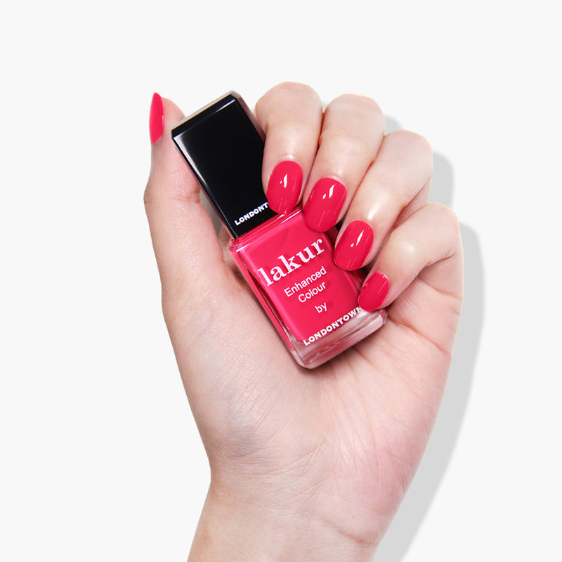 A hand with manicured nails holds a bottle of pink Lakur nail polish in the shade Guilty Pleasure by LONDONTOWN against a white background.