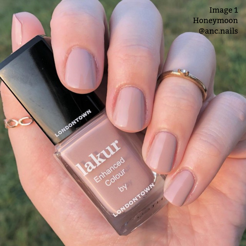 Close-up of a hand holding a bottle of LONDONTOWN's Lakur vegan nail polish in the shade "Honeymoon." The nails are painted in a light pink hue. The person is wearing three gold rings.