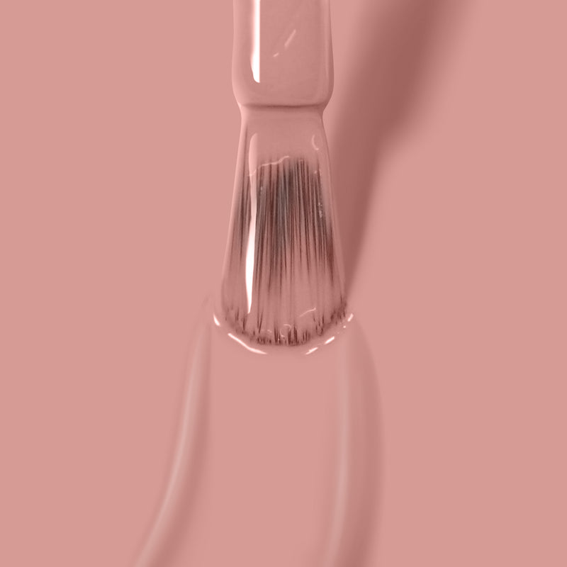 Close-up image of a paintbrush coated in LONDONTOWN Honeymoon light pink glossy vegan nail polish against a matching pink background.