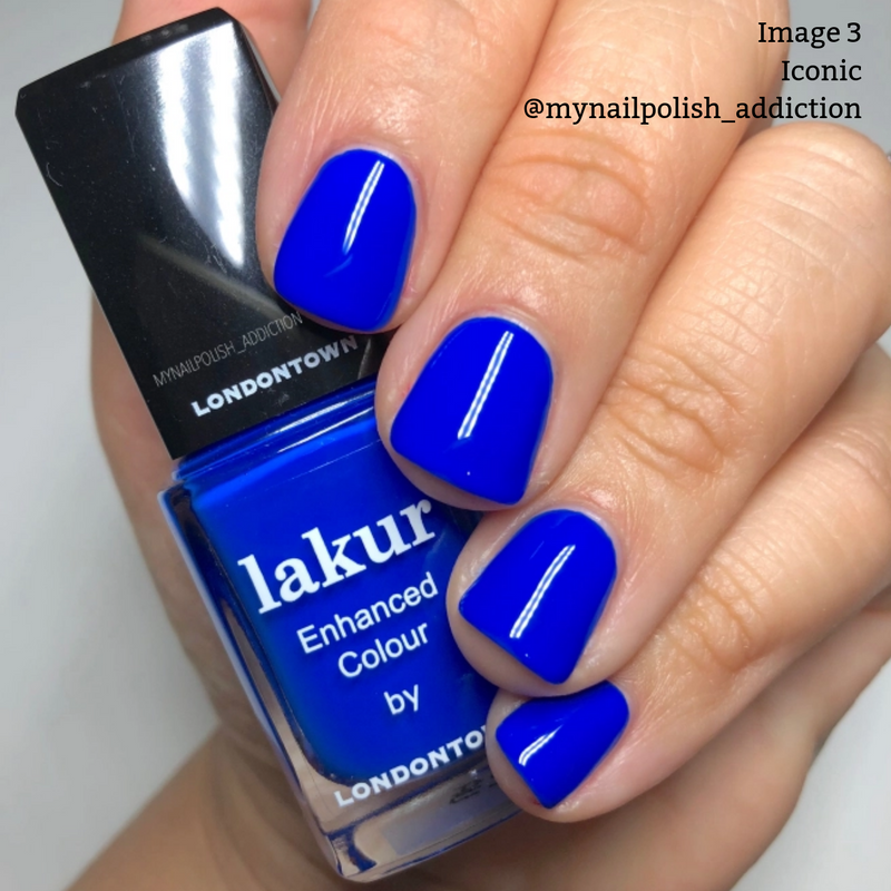 A hand with neatly manicured nails, painted in a vibrant electric blue nail polish, holds a bottle of LONDONTOWN's Lakur Enhanced Colour in the shade "Iconic." This vegan, cruelty-free formula earned the 2019 Allure Best of Beauty Award.