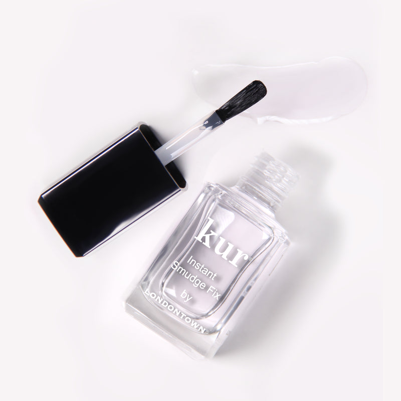 A clear nail polish bottle, labeled "Instant Smudge Fix by LONDONTOWN," is displayed with an open black cap and brush. The visible vegan nail polish liquid on the brush and spread near the bottle makes it ideal for quick nail smudge repairs.