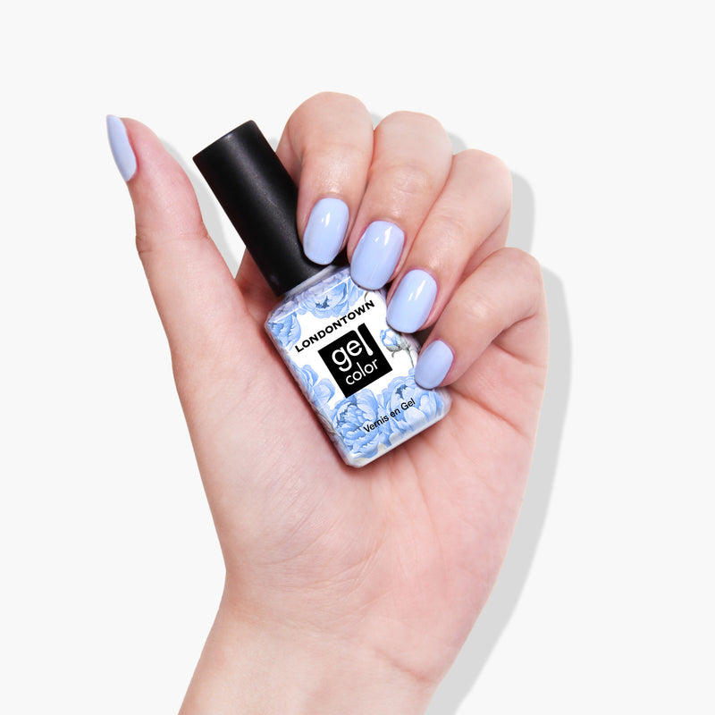 A hand with baby blue skies painted nails holds a bottle of LONDONTOWN's In the Clouds Gel Color nail polish in a matching light blue shade, ready to shine under UV/LED light.