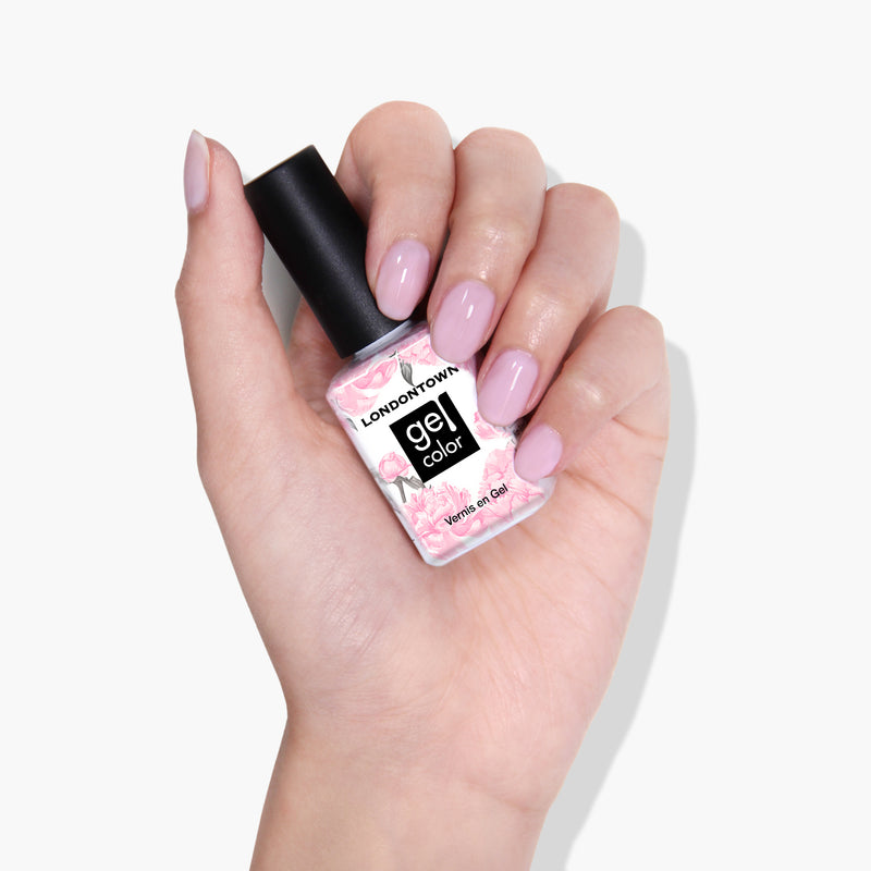 A hand with soft sheer pink polished nails holds a bottle of LONDONTOWN's Invisible Crown gel nail polish in a floral-patterned container against a white background, ready to be set under UV/LED light.