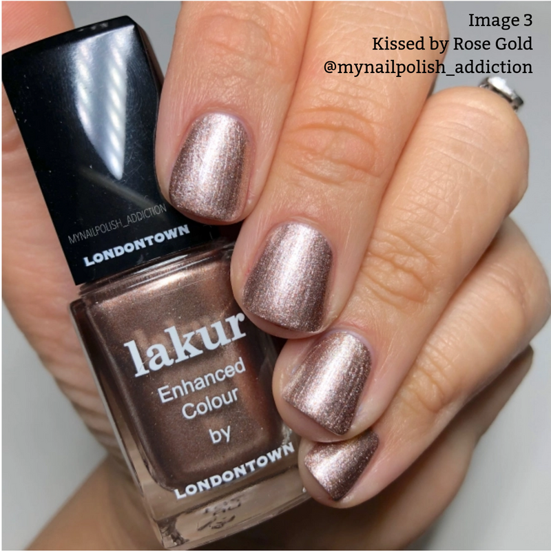 Close-up of a hand with short nails painted in a metallic rose gold color. The hand holds a bottle of LONDONTOWN's Lakur in the shade "Kissed by Rose Gold," known for its cruelty-free and vegan formula. Text reads "Kissed by Rose Gold @mynailpolish_addiction.
