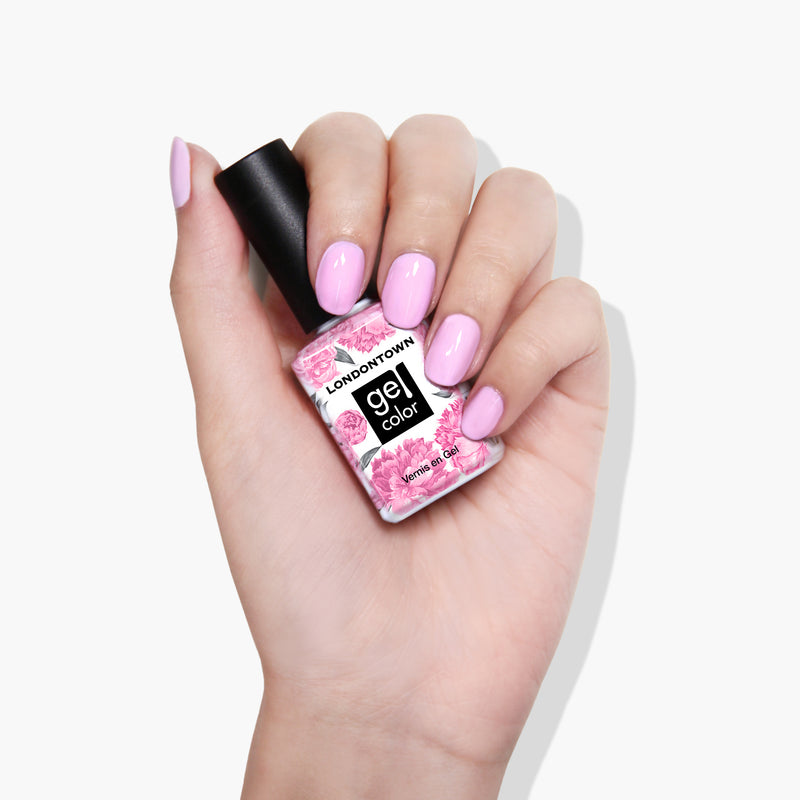 A hand with nails painted in a refreshing muted pink holds a bottle of LONDONTOWN's Lemonade Pop gel color nail polish, beautifully adorned with pink flowers.