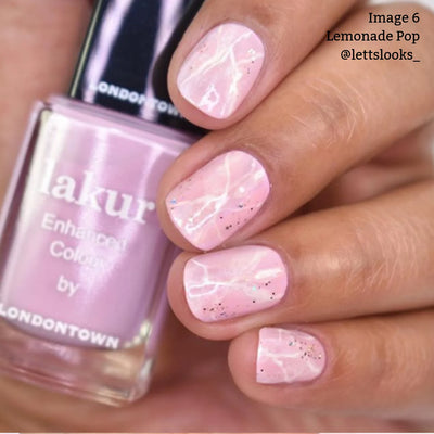 A hand with short, light pink nails featuring a delicate white marble pattern and subtle glitter. A bottle of "Lemonade Pop (Professional)" nail polish by LONDONTOWN, known for its muted pink hue and being vegan cruelty-free, is held in the background.