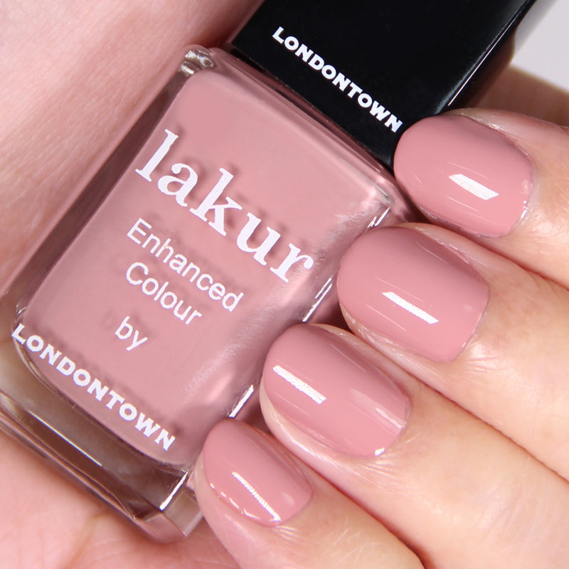 Close-up of a hand showing fingernails painted with a beautiful dusty rose nail polish. The person holds a bottle of LONDONTOWN Lakur Enhanced Colour soft crème nail polish in the shade Mauve Over, which is vegan and cruelty-free.