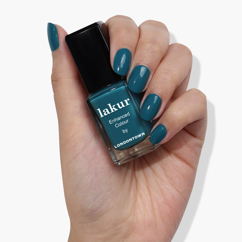 A hand with peacock teal-colored nails holds a bottle of LONDONTOWN's Magpie nail polish in a matching shade against a white background, making a sophisticated statement.