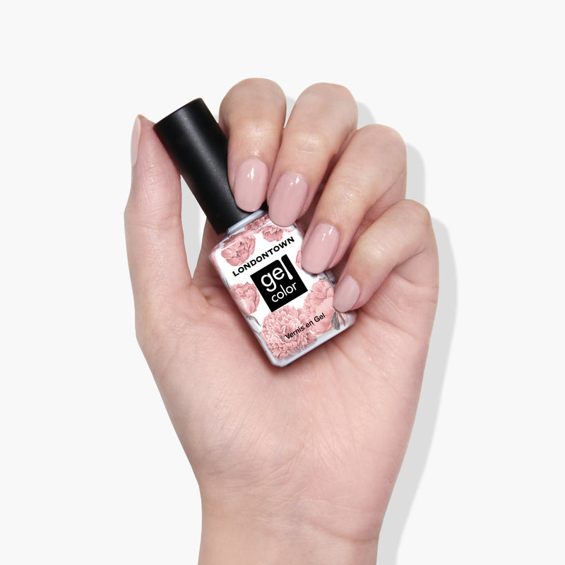 Hand holding a bottle of LONDONTOWN Make Me Blush gel color nail polish with a floral label design. Nails are painted in a light pink shade, showcasing the vibrant, vegan-friendly formula that cures perfectly under UV/LED light.