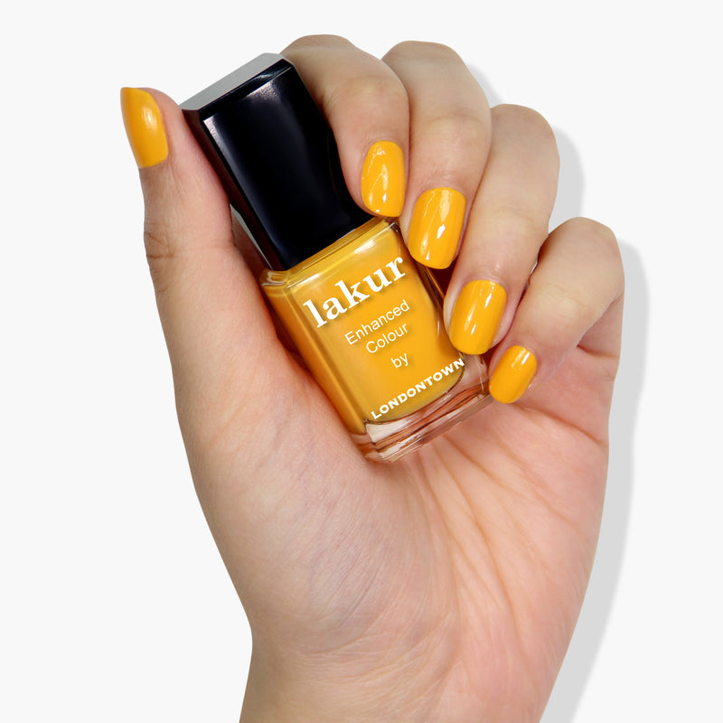 A hand with neatly manicured nails painted yellow holds a bottle of LONDONTOWN's Lakur nail polish in the shade Mango, featuring a 16+ free and cruelty-free formula, against a plain white background.