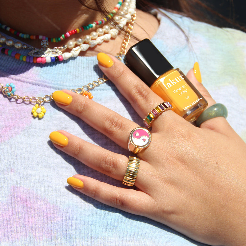A hand with yellow nail polish holds a bottle of LONDONTOWN's Mango, a 16+ free, vegan, and cruelty-free yellow nail polish. The person is adorned with beaded necklaces and several rings, including one featuring a pink yin-yang symbol.