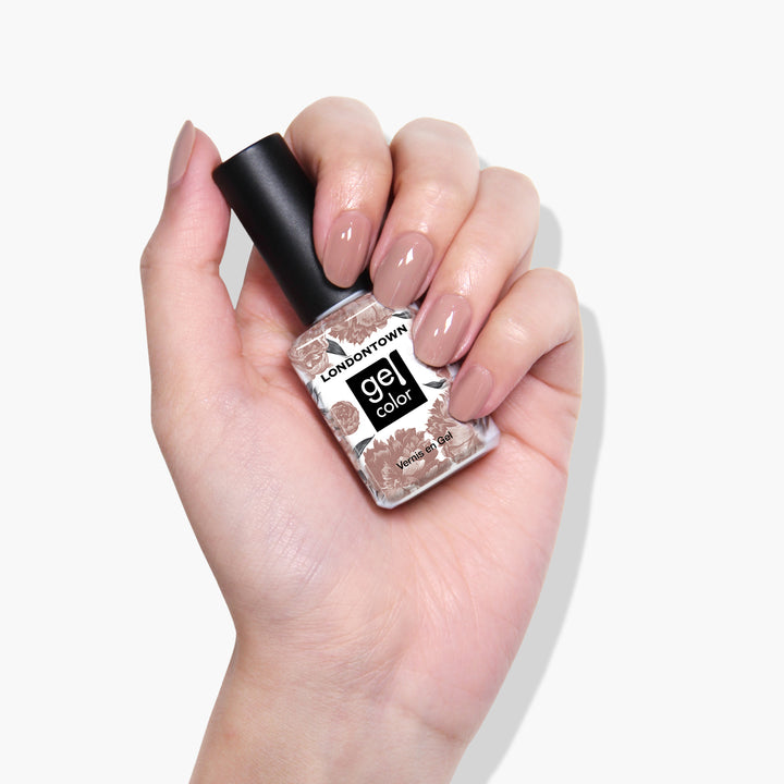 A hand holding a bottle of LONDONTOWN Milk Tea gel nail polish in a deep nude crème shade. The fingernails, polished with the same vegan cruelty-free color, gleam under the light.