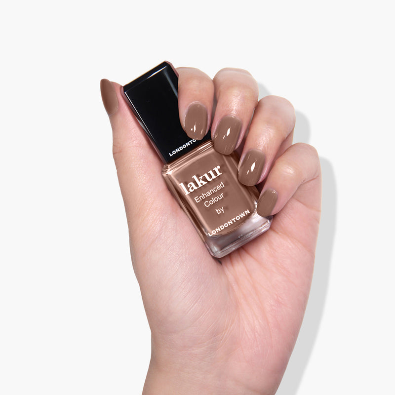 Hand holding a bottle of LONDONTOWN Mudslide nail polish in a neutral deep brown shade, with nails painted in the same color.