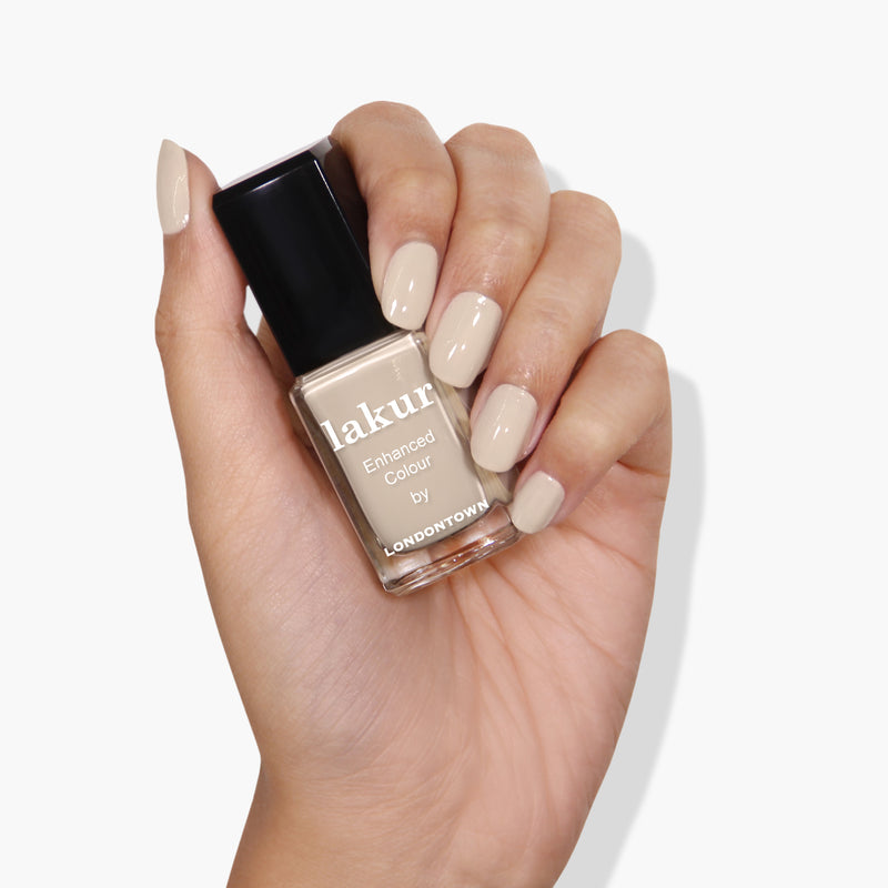 A hand with neatly manicured nails holding a bottle of LONDONTOWN's Lakur Murray Me, a vegan cruelty-free nail polish in a light beige, French vanilla cream color.