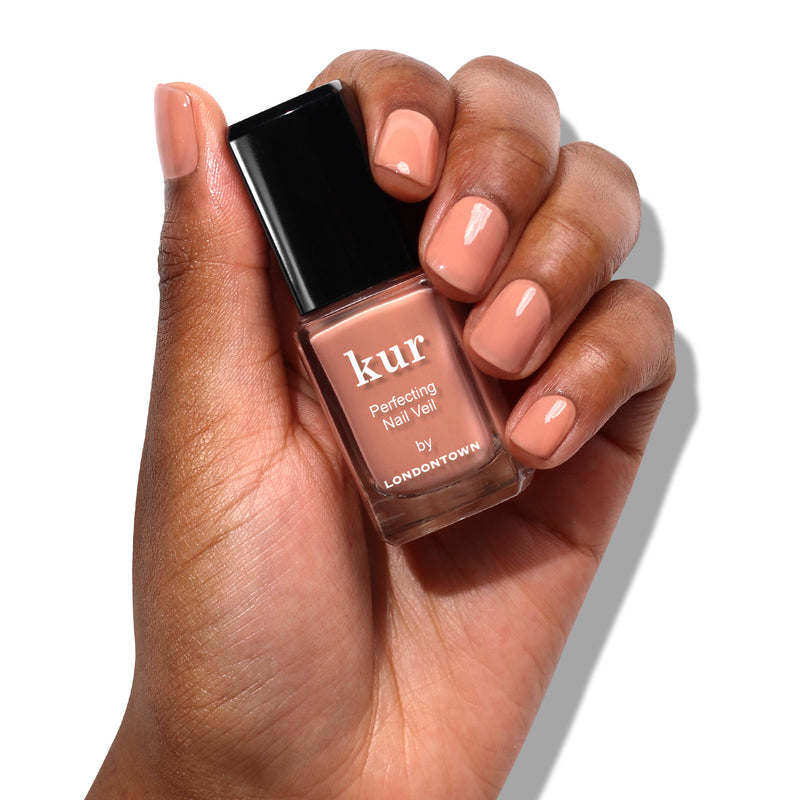 A hand holding a bottle of LONDONTOWN Perfecting Nail Veil #5 nail polish, showcasing nails painted in a matching nude shade. This power-packed formula delivers flawless color and care, ensuring your manicure lasts beautifully.