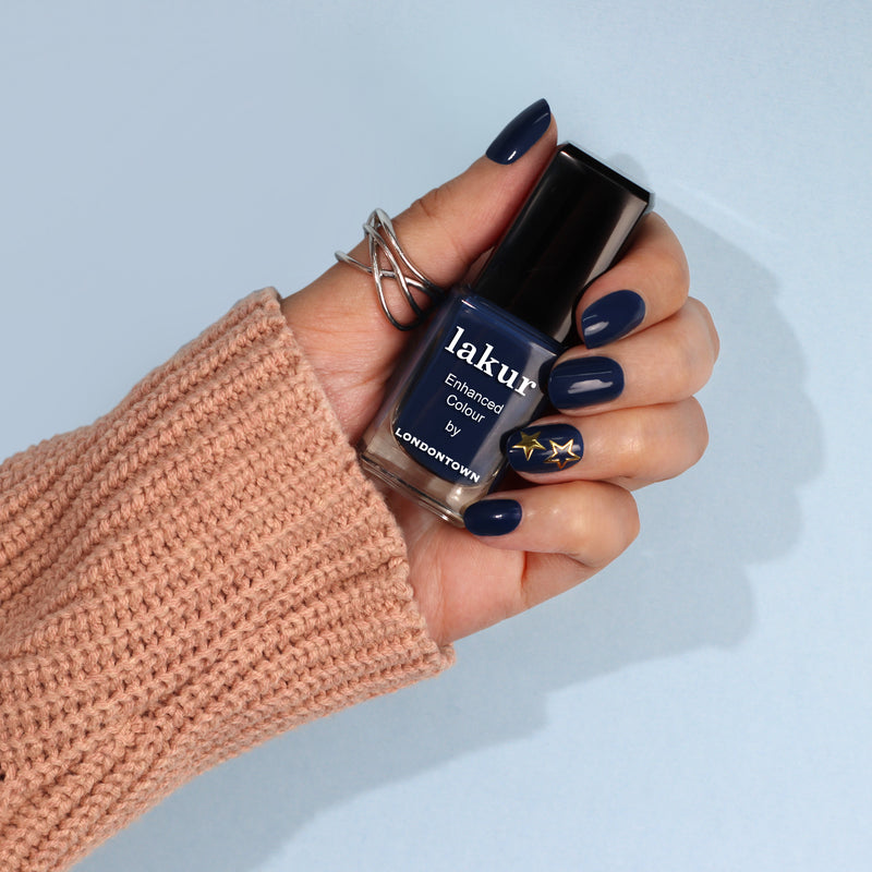 A hand with navy blue painted nails showcases a bottle of Nail Decals - Starbright by LONDONTOWN against a light blue background. The hand, adorned with silver rings and a beige knit sleeve, embodies chic simplicity—perfect for easy nail art enthusiasts.