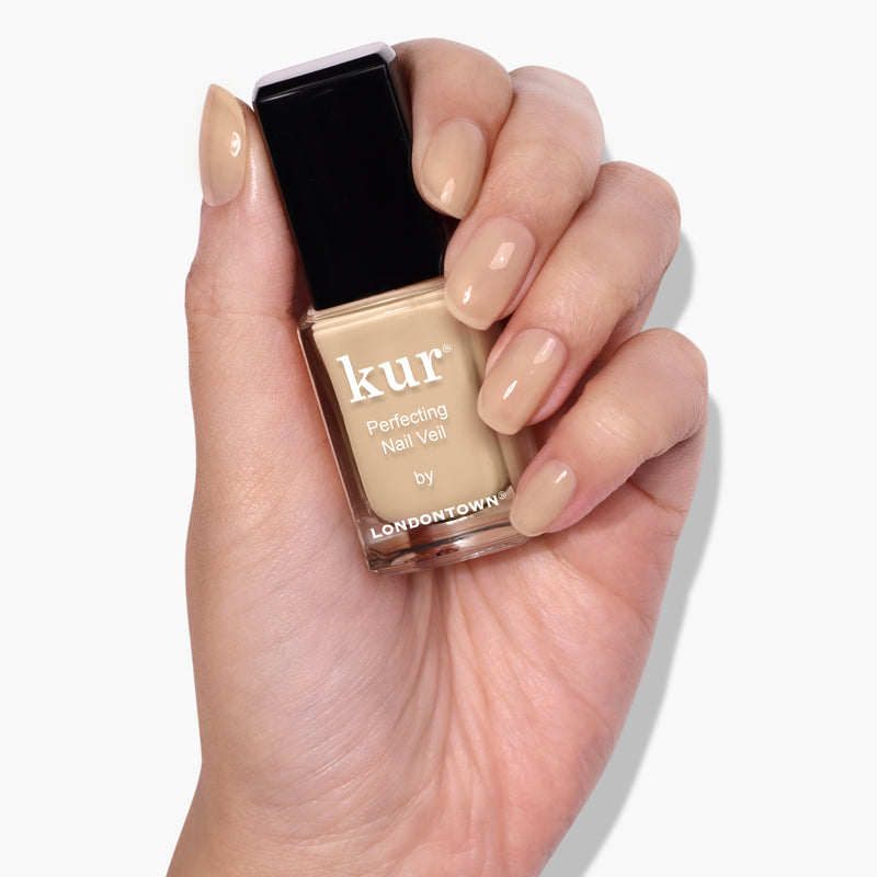 A hand with manicured nails holds a bottle of LONDONTOWN’s Perfecting Nail Veil #2 in a beige shade, offering a hint of color and powerful care.