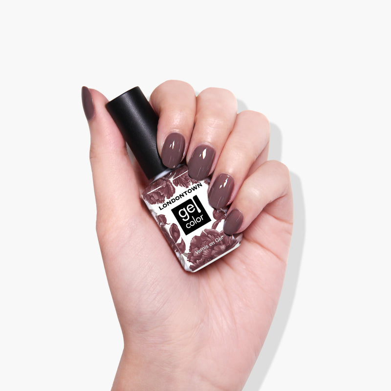 A hand with manicured nails holding a bottle of LONDONTOWN's Natural Charm gel color nail polish in a creamy mauve purple shade against a white background.