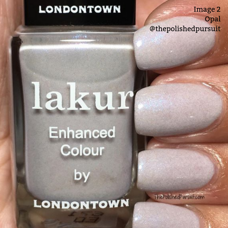 Close-up of a hand with pearlescent white nail polish holds a bottle of LONDONTOWN's "lakur Enhanced Colour" vegan nail polish in the shade "Opal," featuring a hint of shimmering lilac.