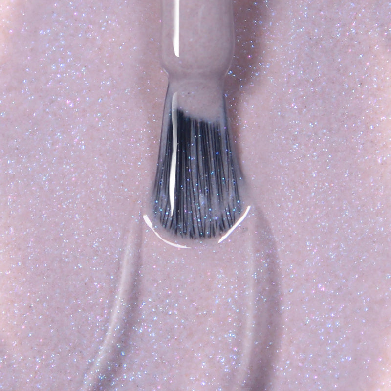 A close-up view of a nail polish brush applying LONDONTOWN's Opal gel color, a light pink glittery shade, on a surface under UV/LED light.