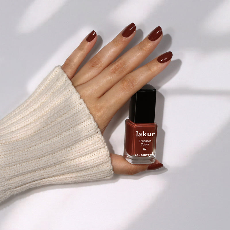 A hand with painted nails showcases a bottle of LONDONTOWN's Oud Mood nail polish against a neutral background. The person is wearing a cream-colored, ribbed sleeve that evokes the cozy warmth of hot chocolate.