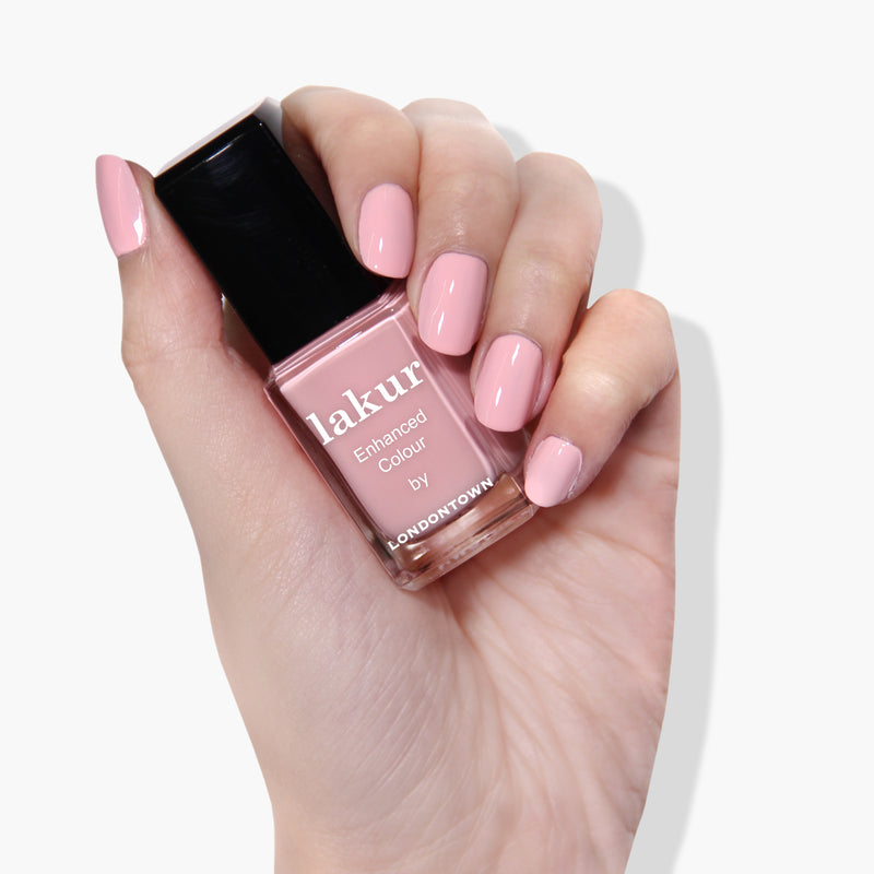 A hand with manicured nails holds a bottle of pastel pink nail polish labeled "Out of Office by LONDONTOWN," featuring the Florium Complex for a superior finish. This vegan, cruelty-free formula ensures your nails look perfect.