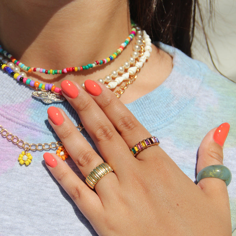 Close-up image of a person wearing colorful beaded necklaces, various gold rings, and Papaya creamy coral vegan nail polish by LONDONTOWN, with a light blue top.