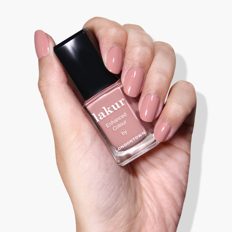 A hand with neatly manicured nails holds a bottle of Lakur nail polish by LONDONTOWN in a pink Pink Sands shade, infused with Florium Complex, against a white background.