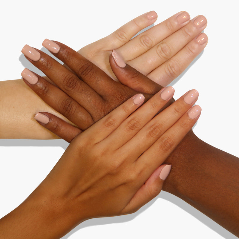 Four hands of different skin tones are layered over each other, all displaying painted nails in a soft nude color reminiscent of ballet slippers. The background is plain white, highlighting LONDONTOWN's vegan and cruelty-free nail polish named Plié.