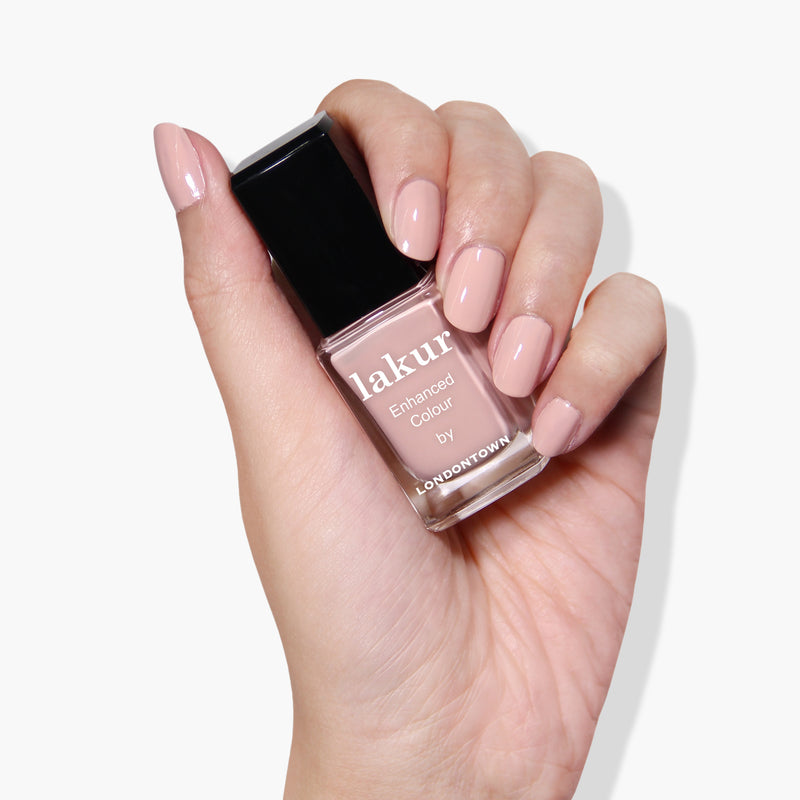 A hand with neatly manicured nails in a subtle ballet slipper shade holds a bottle of Plié by LONDONTOWN, known for its vegan and cruelty-free formula.