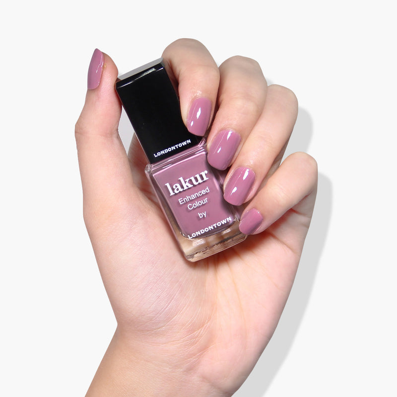 A hand holding a bottle of LONDONTOWN's Plush lakur enhanced colour nail polish in a vegan, cruelty-free mauve crème shade, with nails painted in the same color.