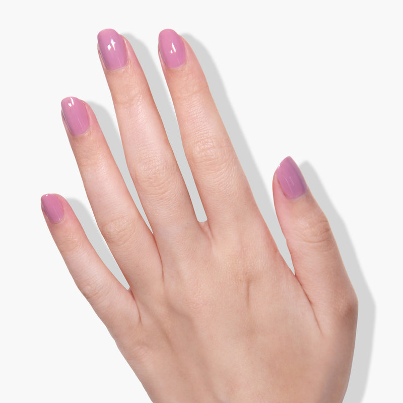 A hand with nails painted in LONDONTOWN's vegan, cruelty-free "Plush" glossy lavender polish is shown against a light gray background.