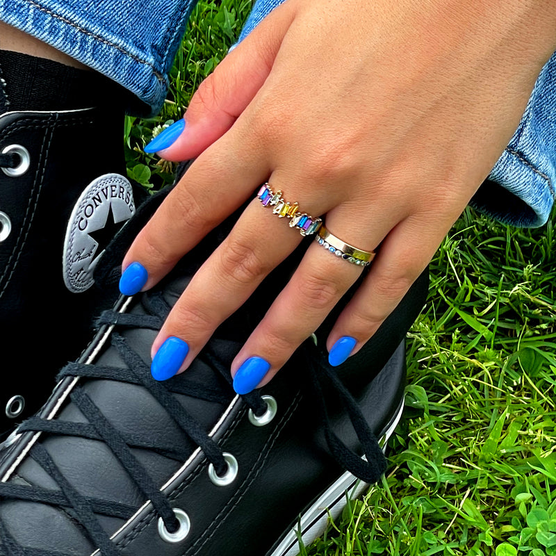 A hand polished with LONDONTOWN's blue, cruelty-free nail lacquer, Poolside Dreams, rests on a black Converse sneaker. The hand is adorned with multiple rings, including one that is multicolored. The person sits on green grass.