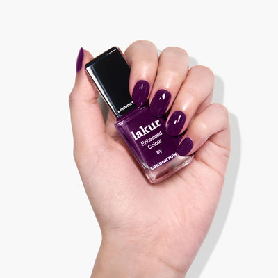 A hand with neatly manicured nails painted in a deep warm plum crème holds a bottle of Portobello Plum (Professional) by LONDONTOWN, a vegan cruelty-free nail polish in the same shade.
