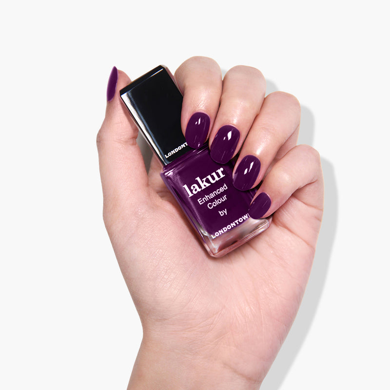A hand with purple-painted nails holds a bottle of Portobello Plum nail polish, a deep warm plum crème by LONDONTOWN, against a white background.