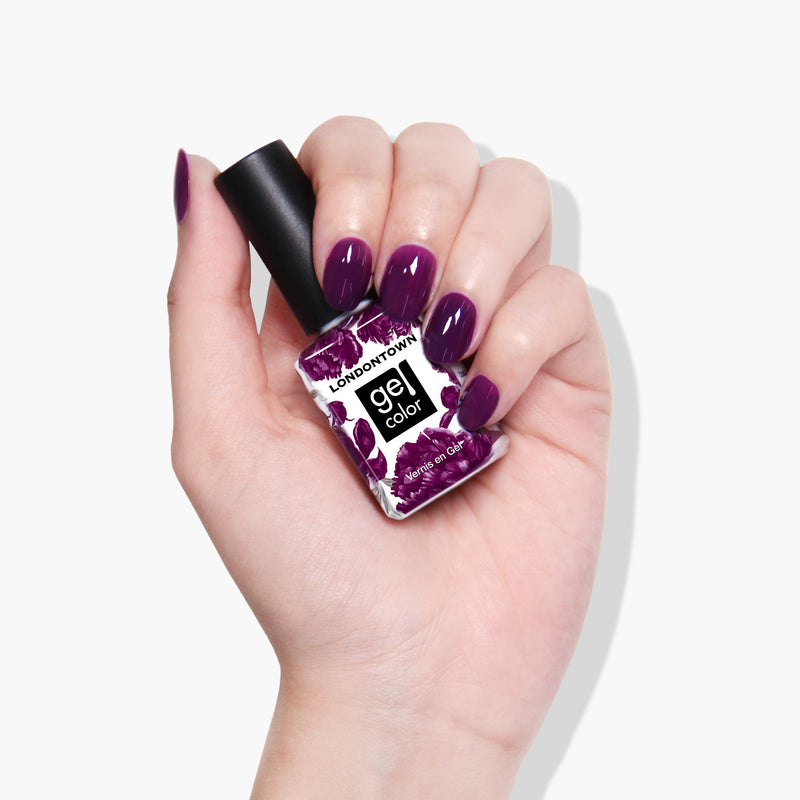 A hand with purple nail polish holds a LONDONTOWN gel color bottle in an elegant shade named Portobello Plum, a deep warm plum crème.