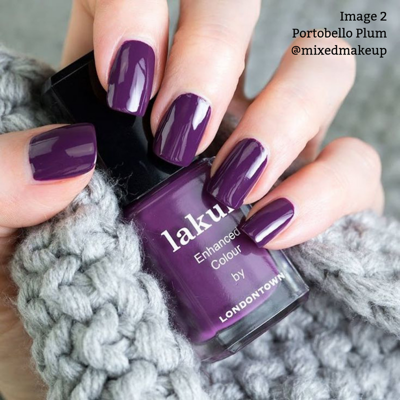 Close-up of hands holding a bottle of LONDONTOWN Portobello Plum nail polish, with nails painted in the same deep warm plum crème, against a grey knit scarf. This vegan and cruelty-free nail polish exudes richness and elegance.