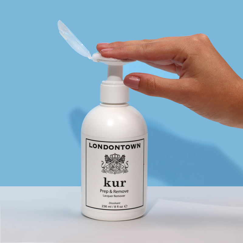 A hand with painted nails presses down on a white LONDONTOWN Lacquer Remover Pump labeled "kur Prep & Remove Lacquer Remover," against a blue background, ensuring no spills for effortless gel removal.