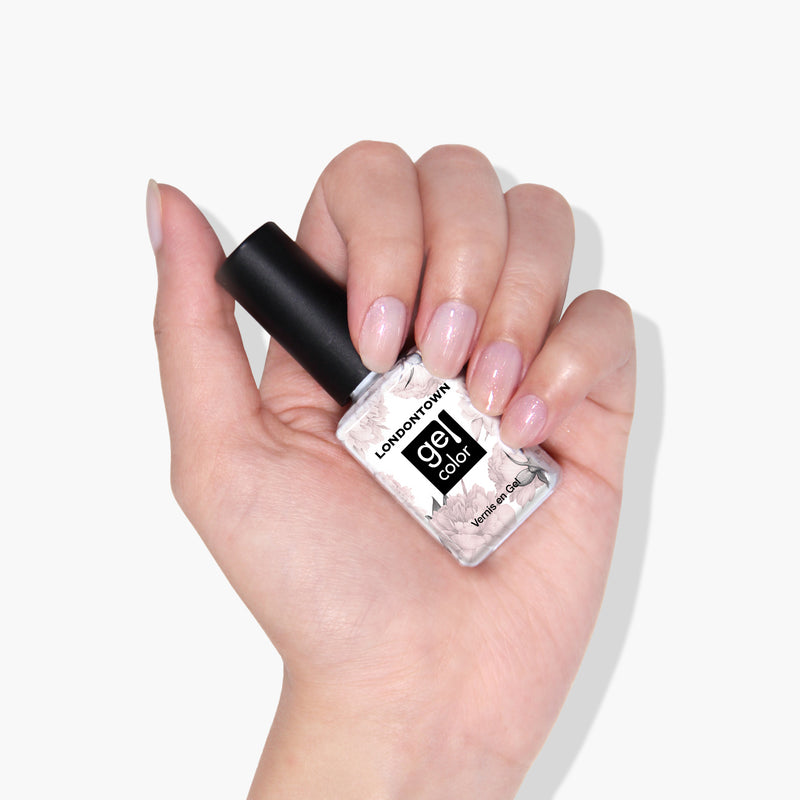 A hand holds a bottle of LONDONTOWN's Princess Awaits gel nail polish, featuring a black cap and a white label, against a white background. The nails are painted with a light, glossy polish that is vegan and cruelty-free.