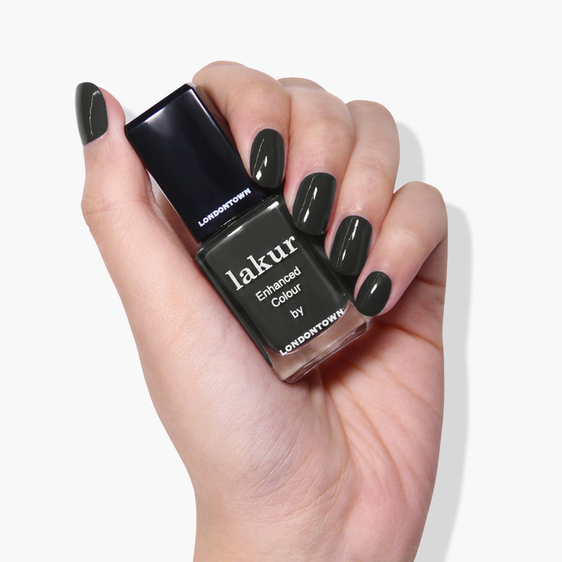 A hand with painted nails holds a bottle of dark green nail polish labeled "Put the Kettle On by LONDONTOWN," a 16+ Free, vegan cruelty-free nail polish sure to make your manicure stand out.