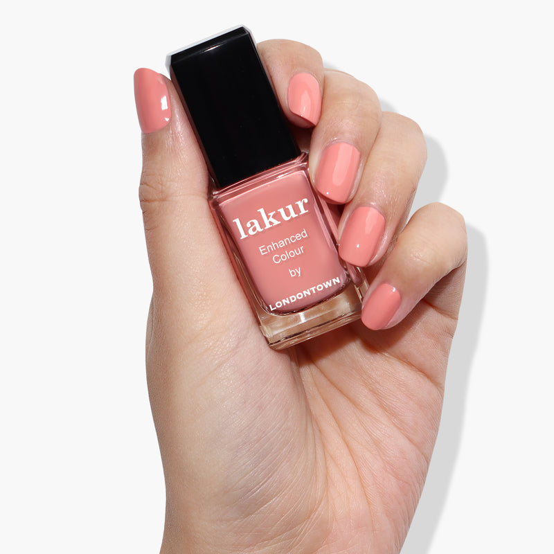 A hand holding a bottle of LONDONTOWN Rose-Tinted Glasses nail polish in a peach shade reminiscent of Himalayan pink sea salt. The nails are painted with the same spring-ready hue.