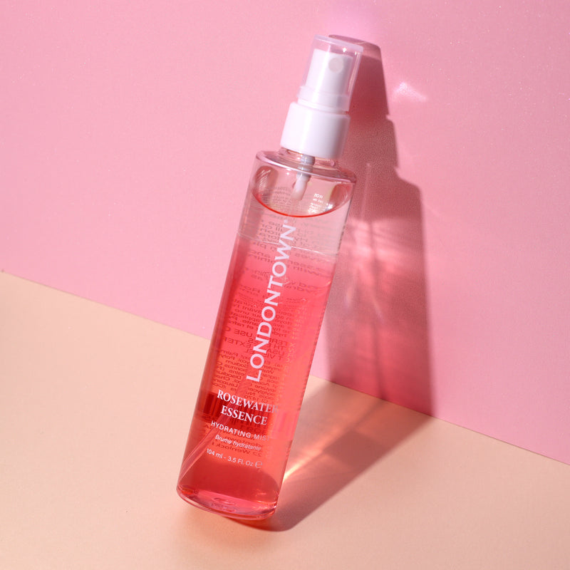 A clear plastic spray bottle labeled "LONDONTOWN Rosewater Essence Facial Mist" stands on a pink and beige background, promising rose-infused hydration.