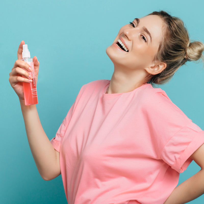 A woman in a pink shirt smiles while holding and spraying a bottle of LONDONTOWN Rosewater Essence Facial Mist, creating a refreshing cloud against a blue background.