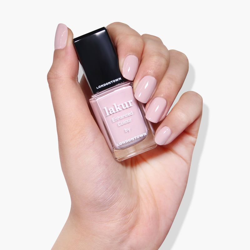 A hand holds a bottle of LONDONTOWN's Rosewater Vegan Enhanced Colour Nail Polish in a light pink shade. The nails are painted with the same color.